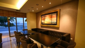 A dining area with a long table and multiple chairs, a large painting on the wall, and a view of the ocean through the windows. - Hotel Flisvos (Kalamata)