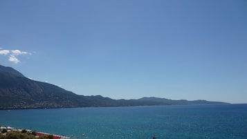 Uma praia costeira com águas azuis cristalinas, montanhas ao fundo e alguns guarda-sóis e cadeiras de praia.