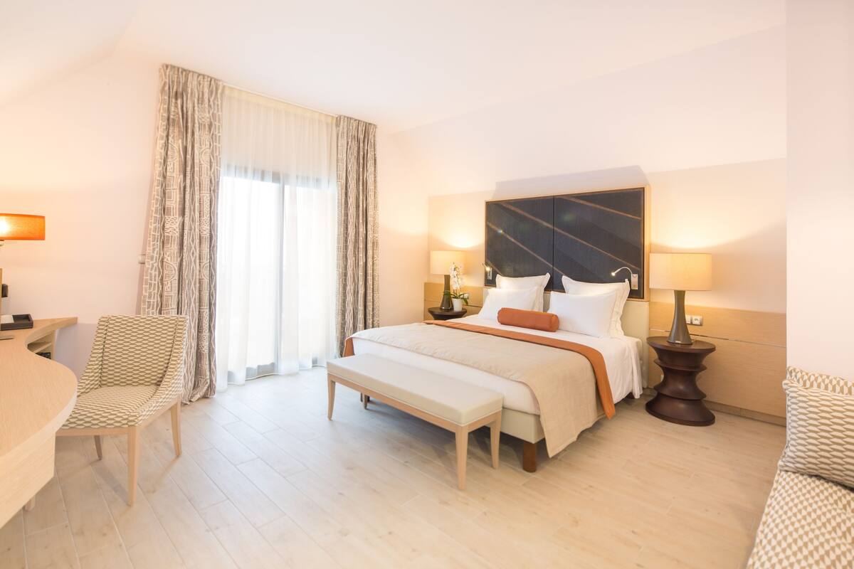 A hotel room with a large bed, a desk, a chair, and a bench.