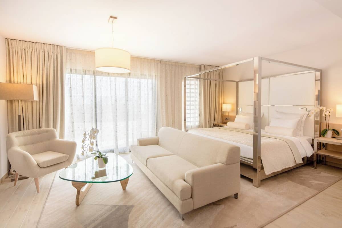 A modern hotel room with a four-poster bed, a sofa, a glass coffee table, and a lamp.
