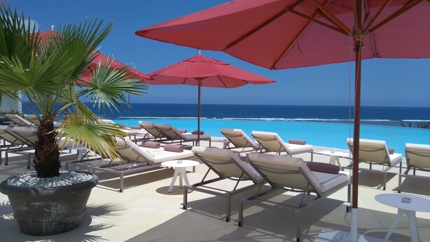Zona junto a la piscina con sillones de descanso y sombrillas rojas, con vista a un océano azul y despejado.