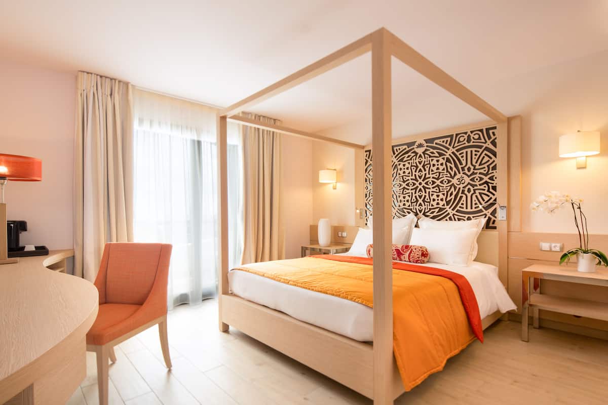 A hotel room with a four-poster bed, a desk with a chair, a nightstand, and a potted plant.