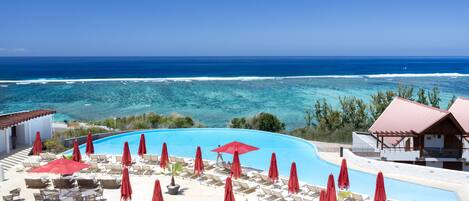 Ein Hotelpool mit roten Sonnenschirmen und Liegestühlen mit Blick auf einen Strand und den Ozean.