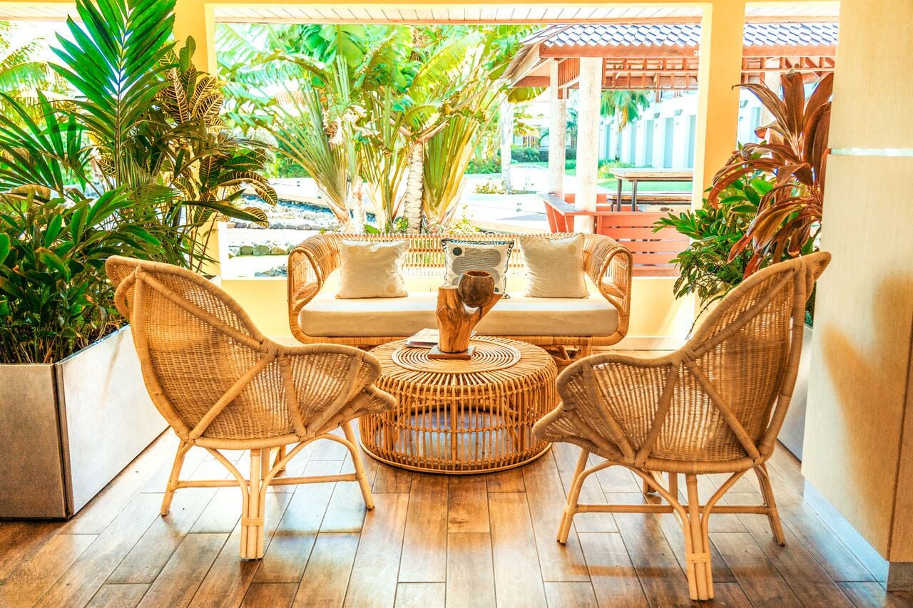 A wicker sofa with cushions and a round table in a sunlit room with wooden flooring and potted plants.