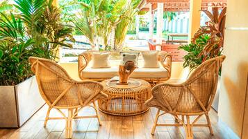 A wicker sofa with cushions and a round table in a sunlit room with wooden flooring and potted plants.