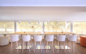 A modern dining area with a long table, white chairs, and a view of palm trees through large windows. - Akoya Hotel & Spa (Saint-Paul)