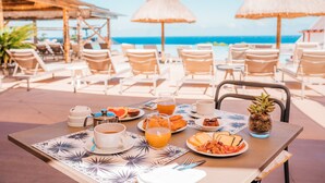 Una mesa dispuesta con alimentos para el desayuno, como cruasanes, queso y fruta, con vista a una playa con sillas de descanso y sombrillas.