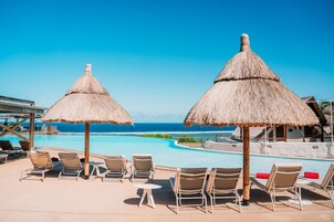 Zona junto a la piscina con sillones de descanso y sombrillas de paja, con vista a un océano azul y cristalino.