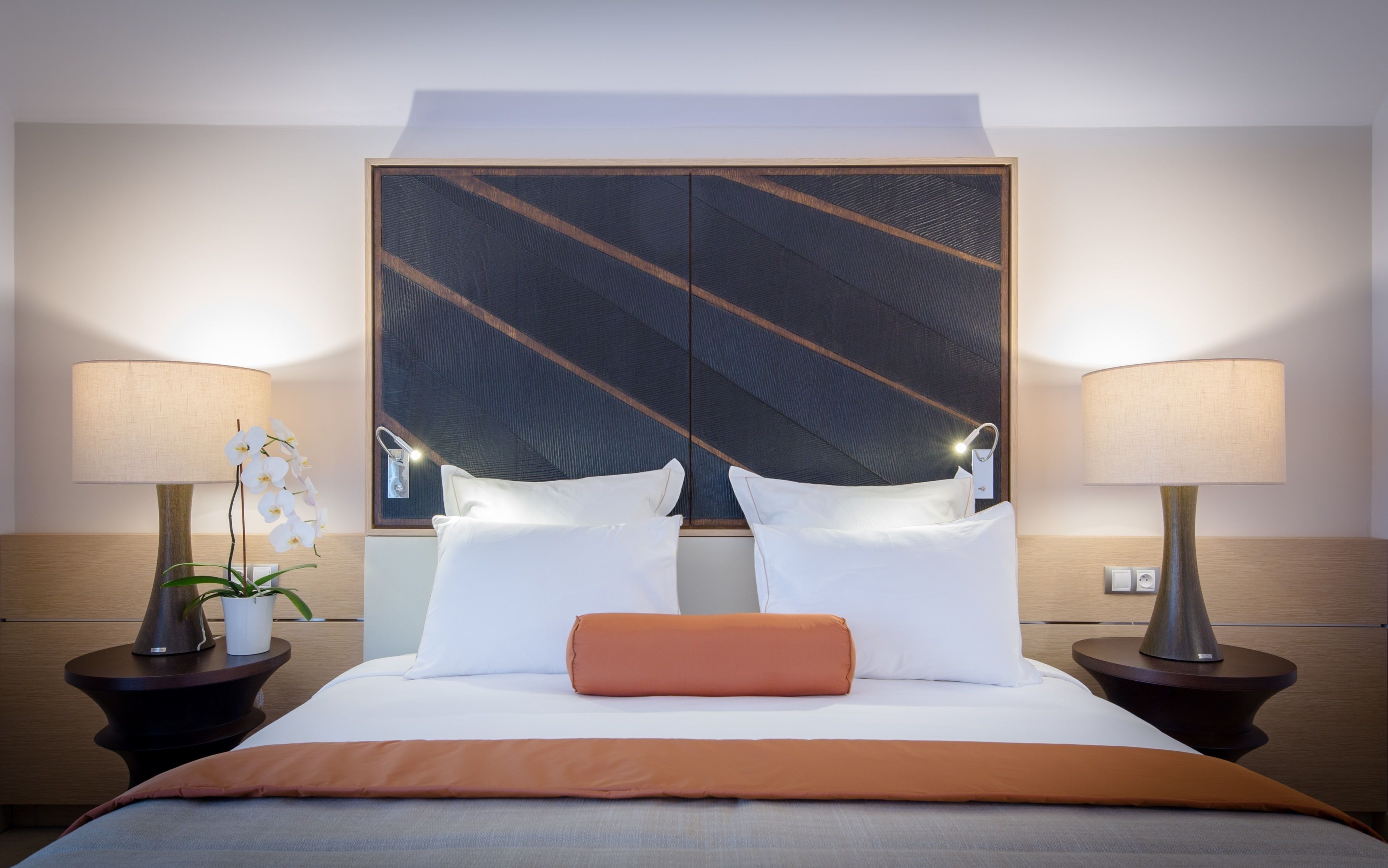 Quarto de hotel com uma cama grande, duas luminárias de cabeceira, uma mesa de cabeceira com uma planta e uma cabeceira decorativa.