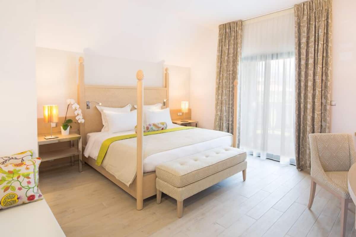A hotel room with a four-poster bed, a desk with a chair, a bedside table with lamps, and a large window with curtains.