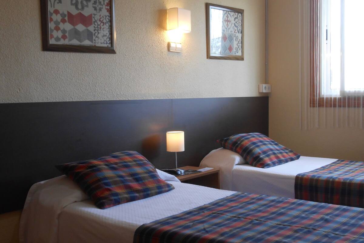 Une chambre d’hôtel avec deux lits, chacun recouvert d’une couverture à carreaux, une table de chevet avec une lampe et deux appliques murales.