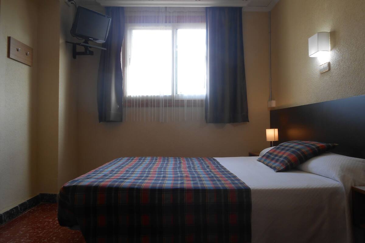 Une chambre d’hôtel avec un lit, un téléviseur fixé au mur, une fenêtre avec des rideaux et une lampe de chevet.