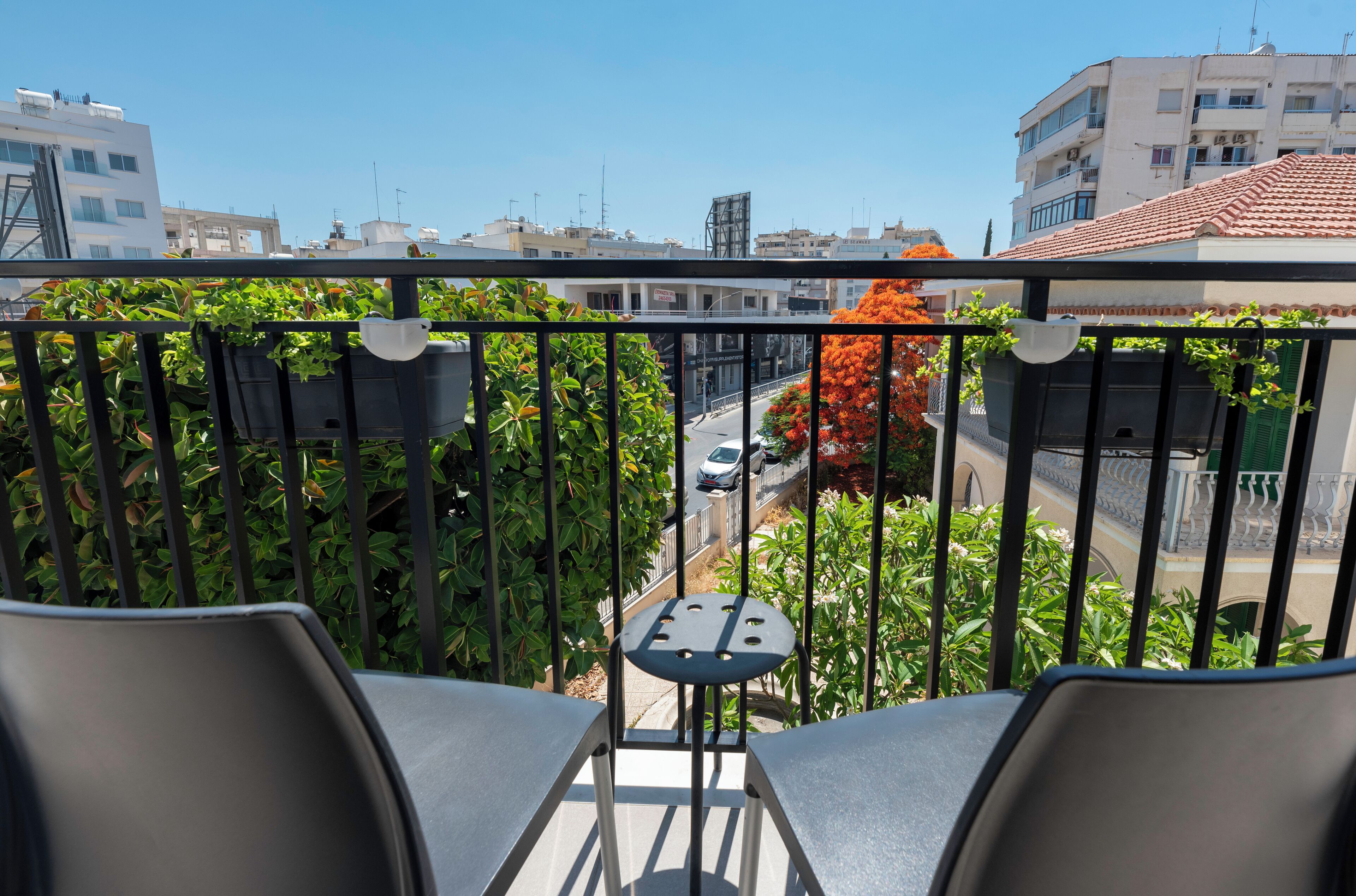 Un balcon avec deux chaises et une petite table, donnant sur une rue bordée d’immeubles et sur un arbre aux fleurs oranges.