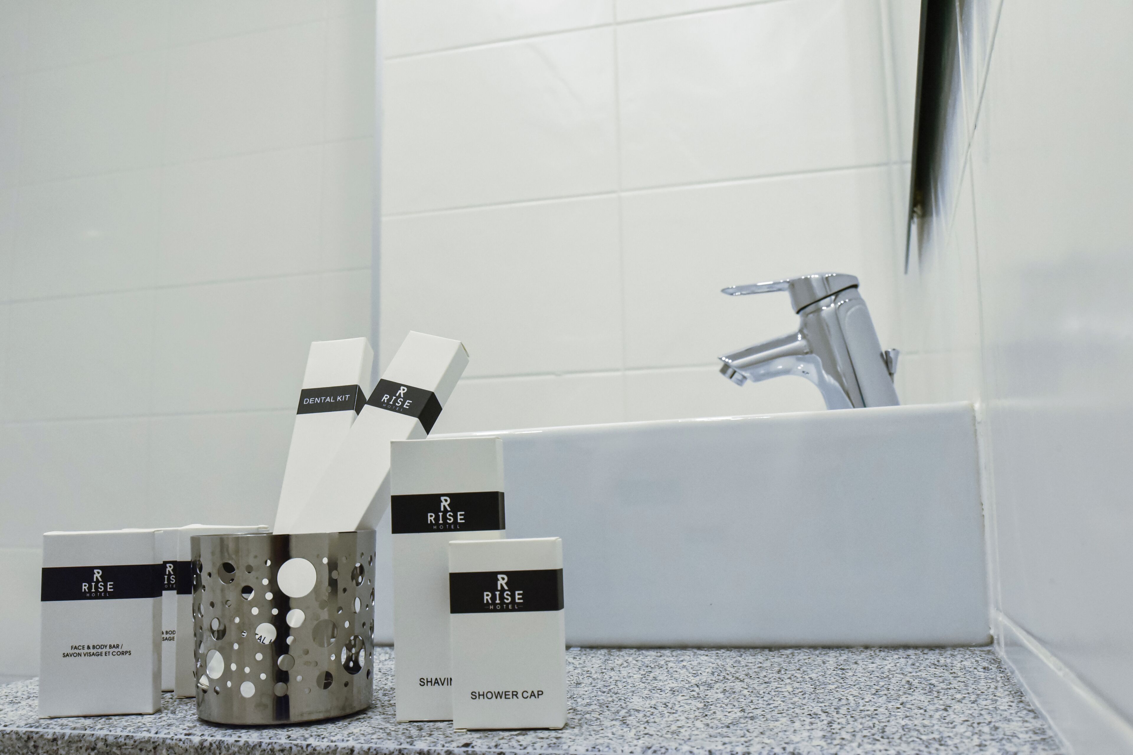 A bathroom with a white sink, a mirror, and a shower area. There are boxes labeled "R Rise Hotel" and "Shower Cap" on the counter.