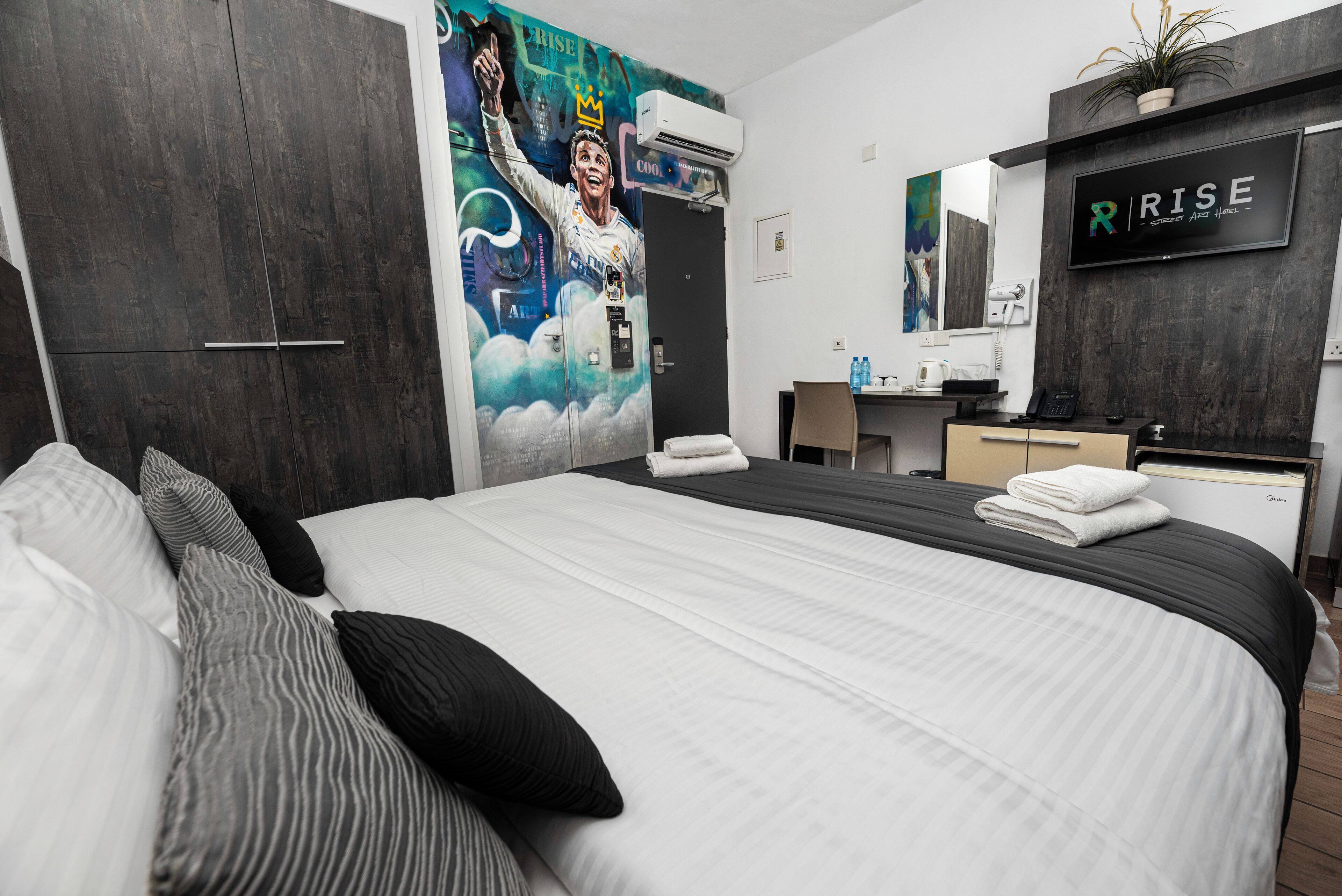 A modern hotel room with a large bed, a wardrobe, a desk with a chair, a mirror, and a TV mounted on the wall.
