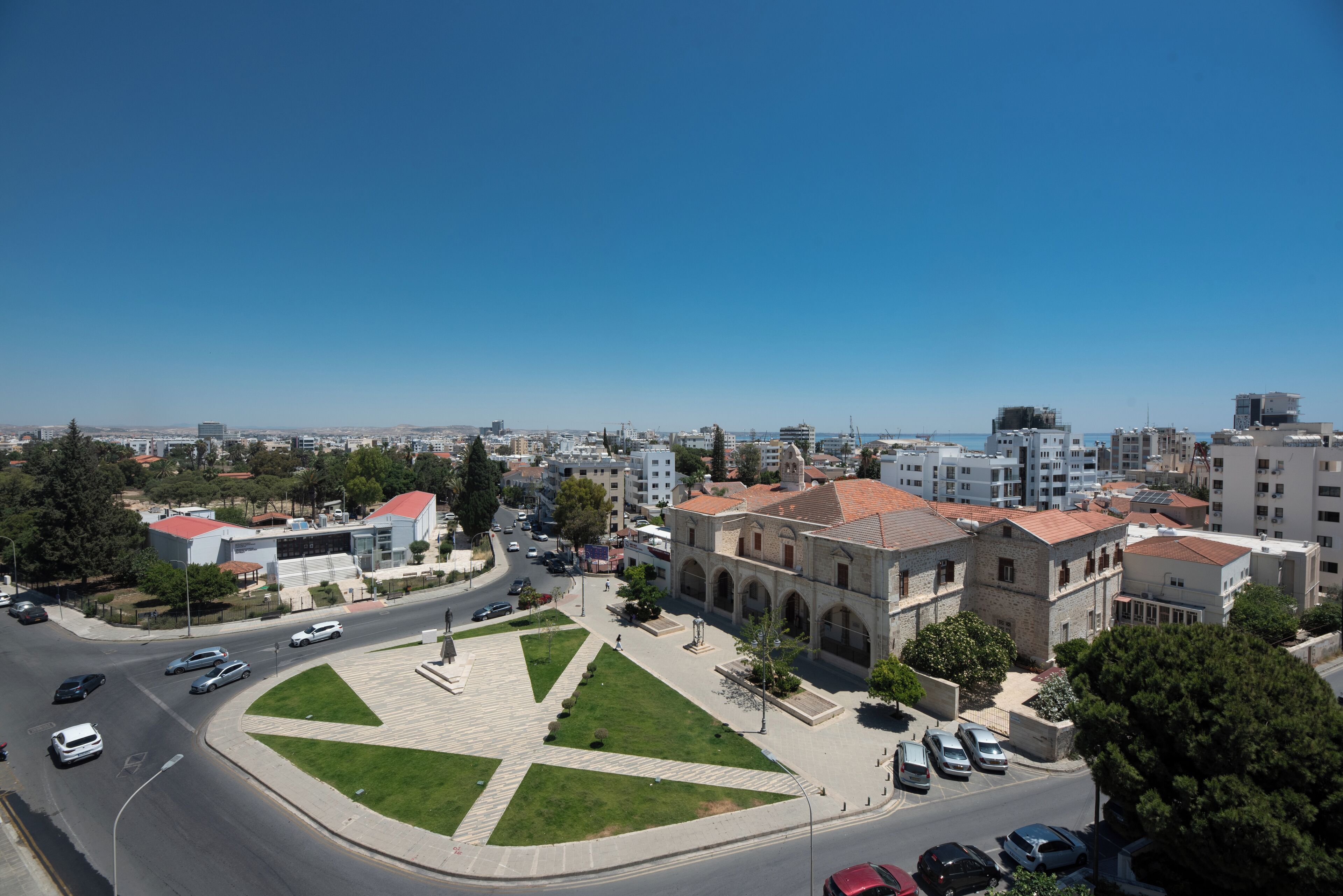 Un paisaje urbano con un edificio destacado, que presenta un techo rojo y ventanas arqueadas, rodeado de un espacio verde cuidado y un área de estacionamiento.