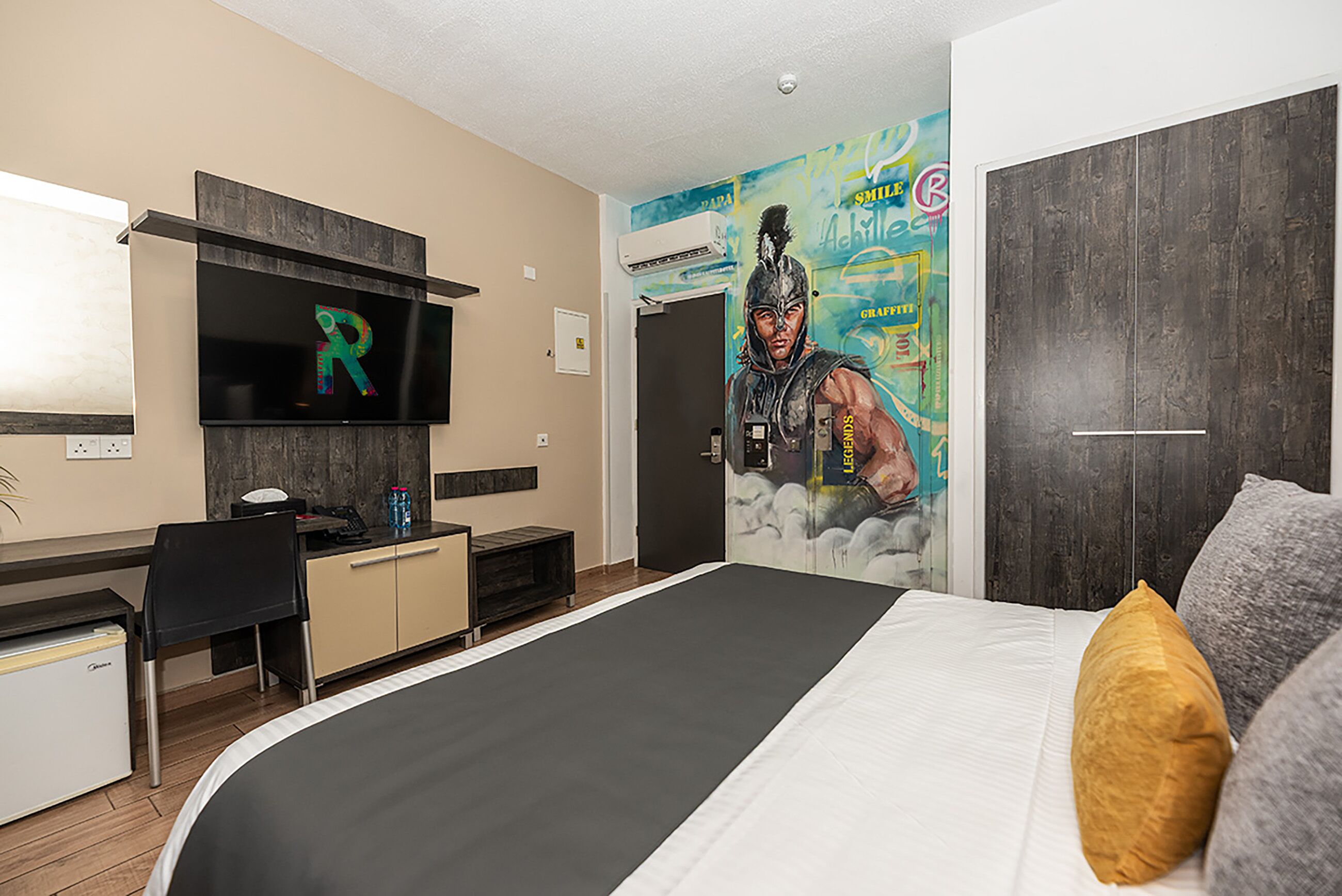 Una camera d'hotel con un letto, una scrivania, una sedia, una televisione e un affresco sulla parete.