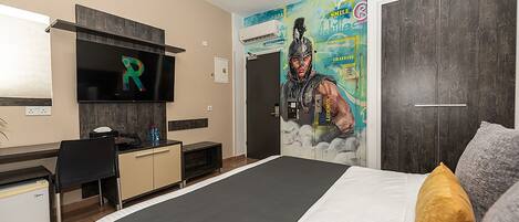 Una camera d'hotel con un letto, una scrivania, una sedia, una televisione e un affresco sulla parete.