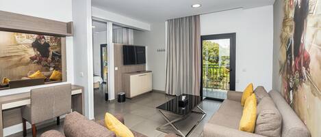 A modern living room with a sofa, a coffee table, and a TV. There is a kitchen area with a counter and a window.