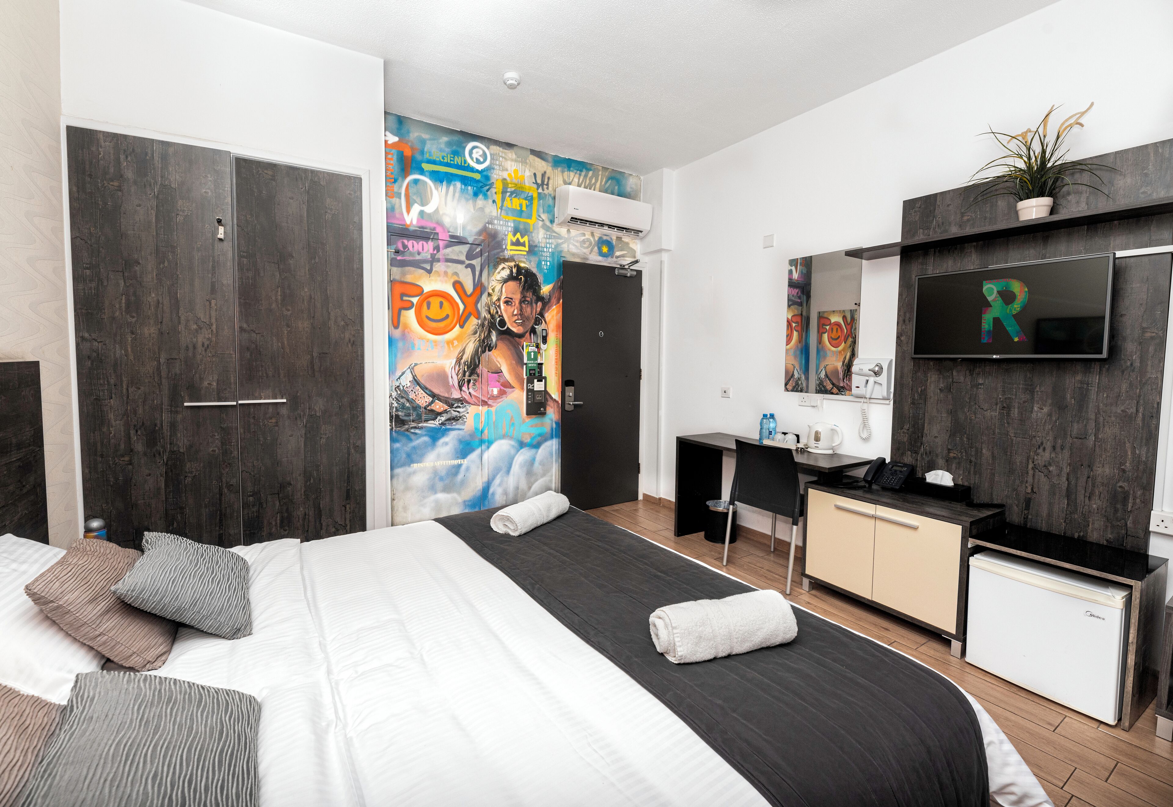 A modern hotel room with a large bed, a desk, a small fridge, and a TV mounted on the wall.