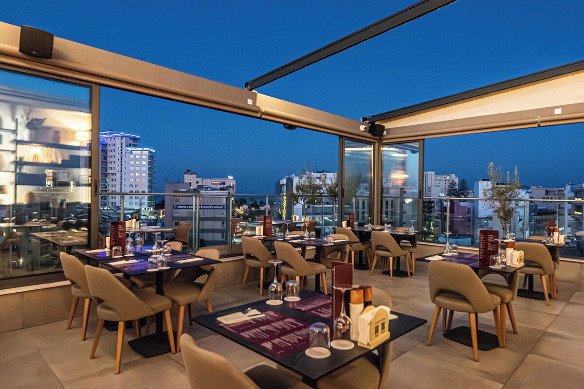 A rooftop dining area with tables and chairs, large windows, and a cityscape view.