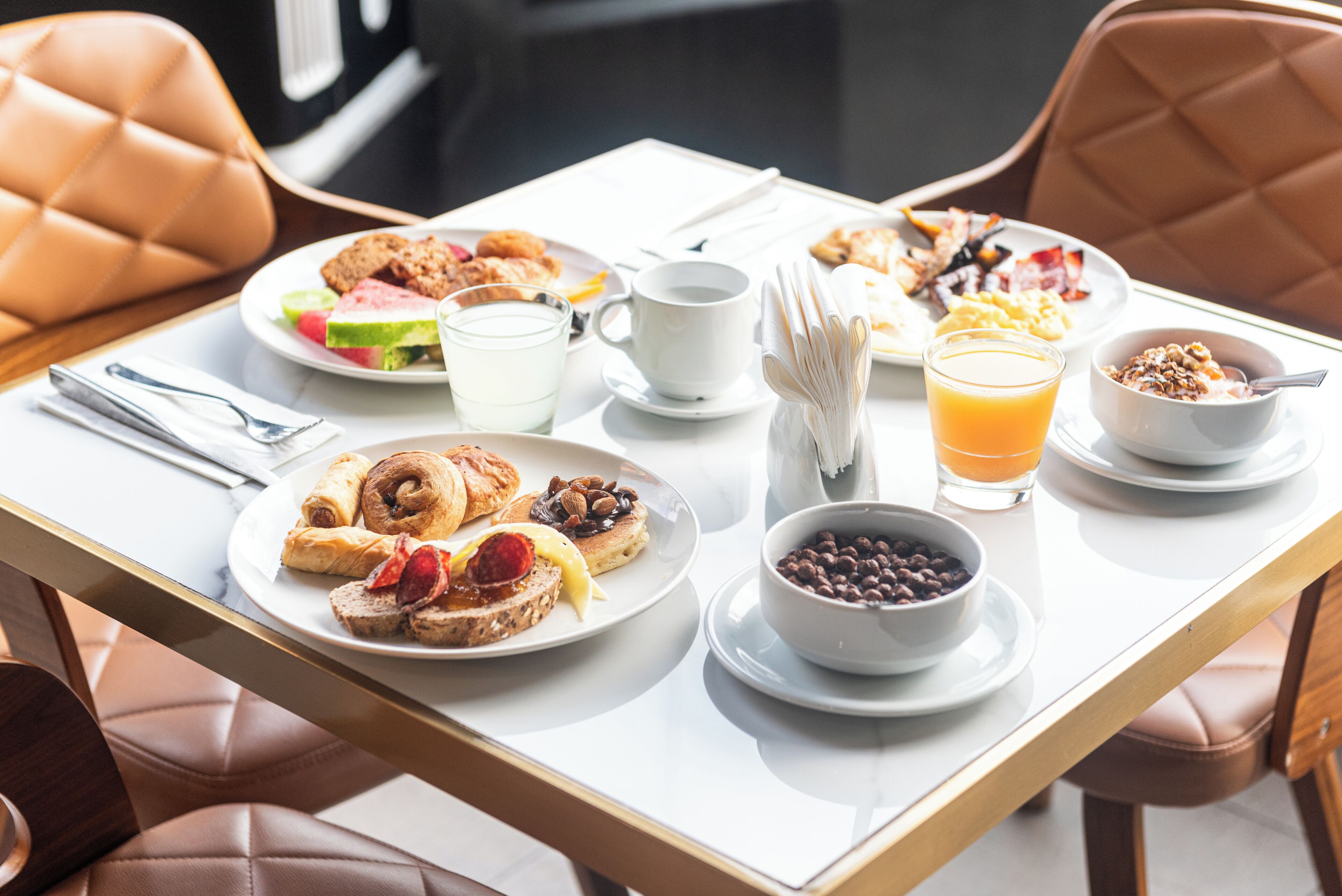 A breakfast table with plates of food, a coffee cup, a glass of orange juice, and a bowl of cereal.