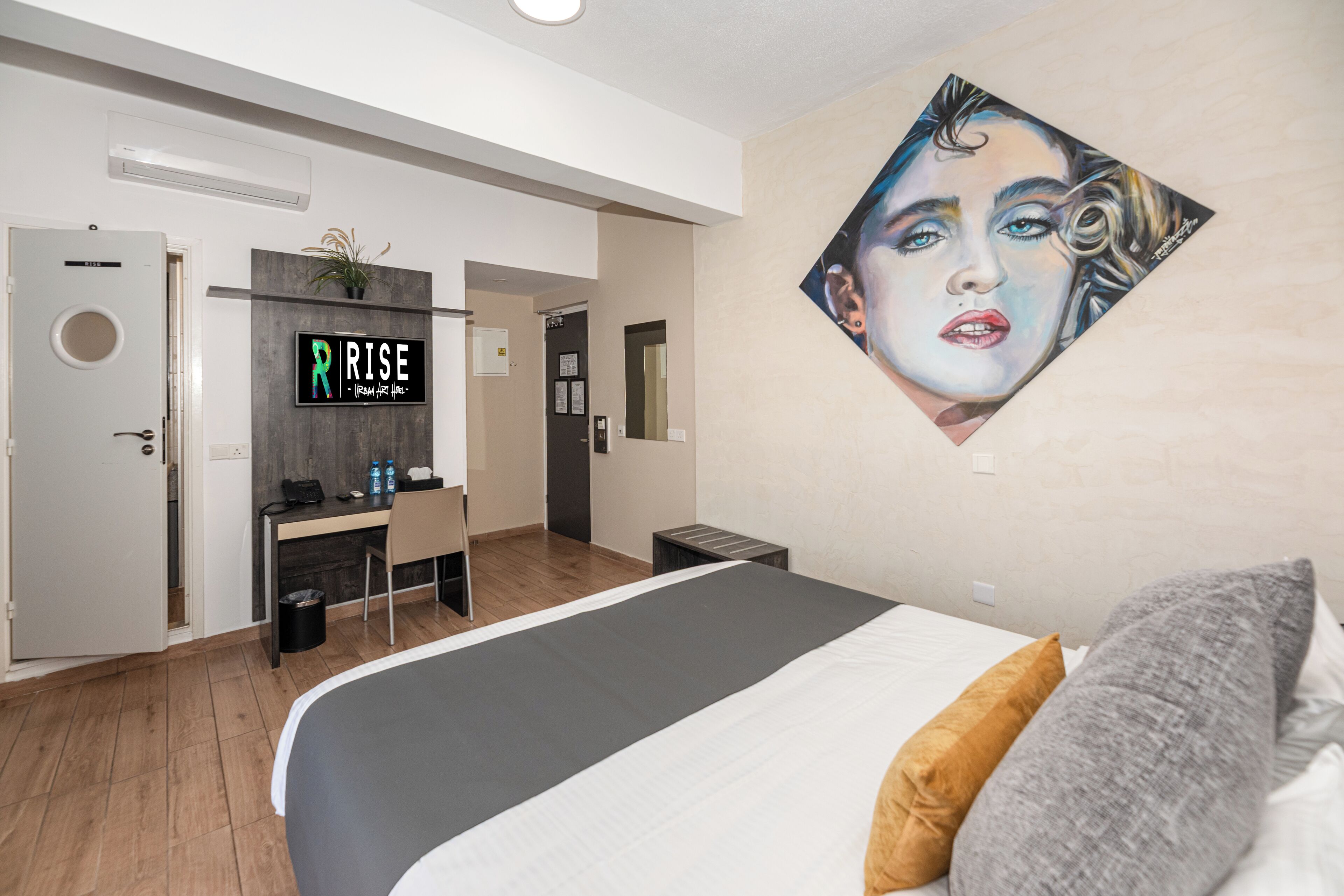 A modern hotel room with a bed, a desk, a chair, a trash bin, and a wall-mounted sign.