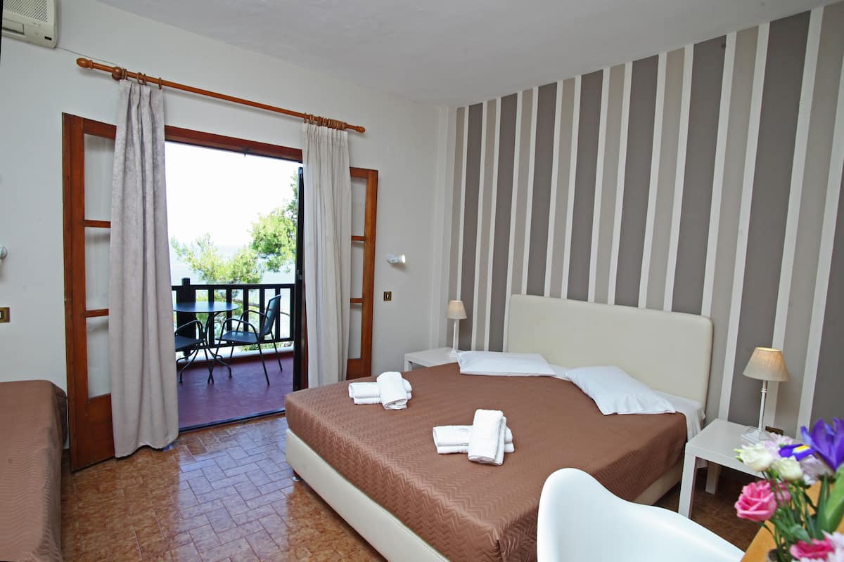 A hotel room with a bed, a bedside table with a lamp, a chair, a balcony with a table and chairs, and a view of greenery outside.