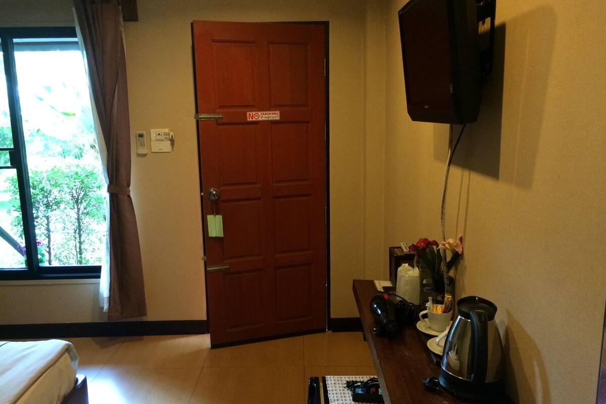 Ein Hotelzimmer mit Holztür, einem an der Wand befestigten Fernseher, einem kleinen Tisch mit Wasserkocher und einem Fenster mit Vorhängen.