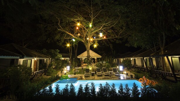 Een nachtzicht van een resort met een zwembad, ligbedden en een met lichtjes versierde boom.