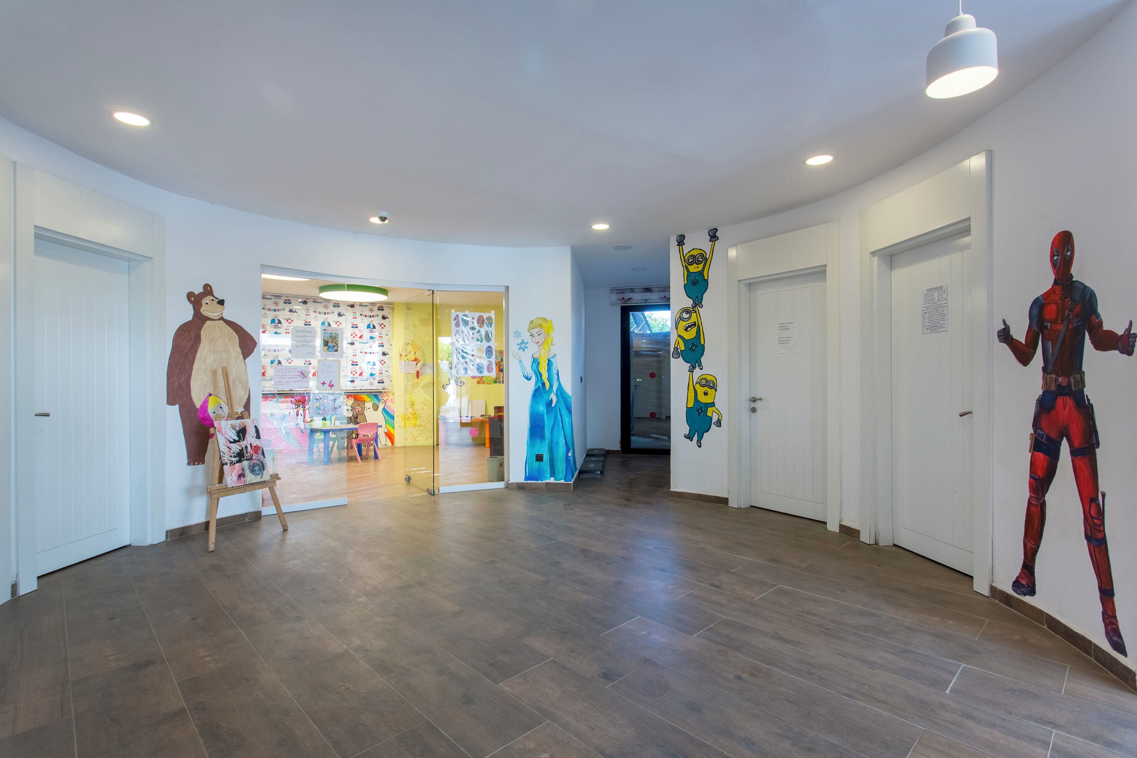 A hallway with wooden flooring, white walls, and doors. There are wall decorations including a bear, a blue dress, and a character from a popular animated film.