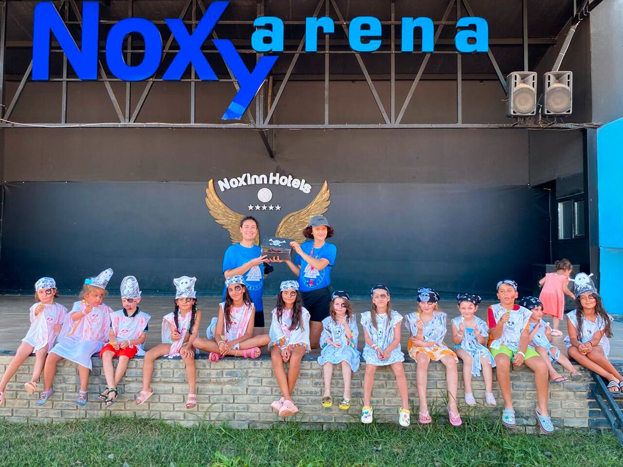 A group of children sitting in front of a building with a sign that reads "Noxy arena" and "NoXInn Hotels".