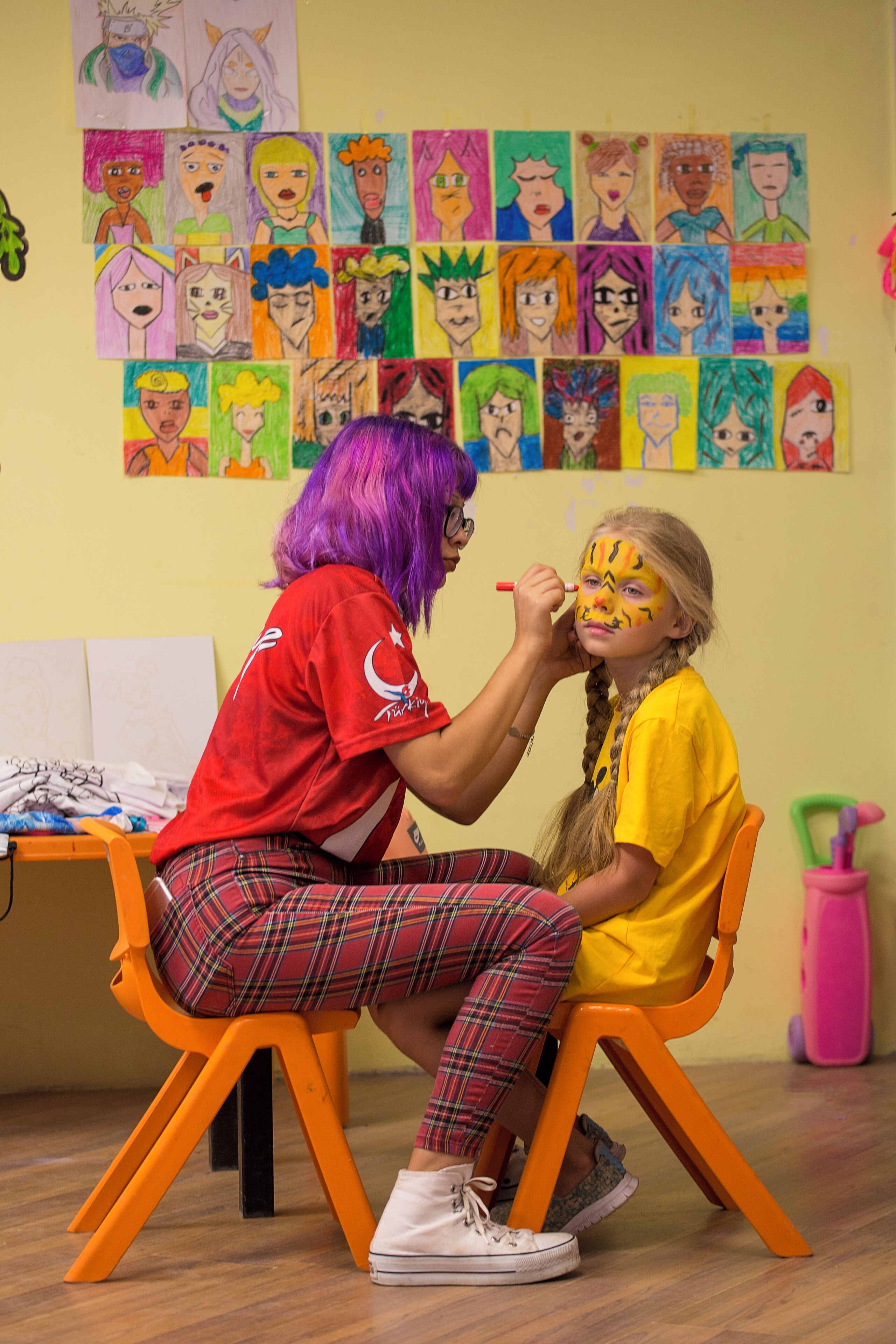 Niño recibiendo pintura en la cara de un adulto en una habitación con un muro de dibujos coloridos.