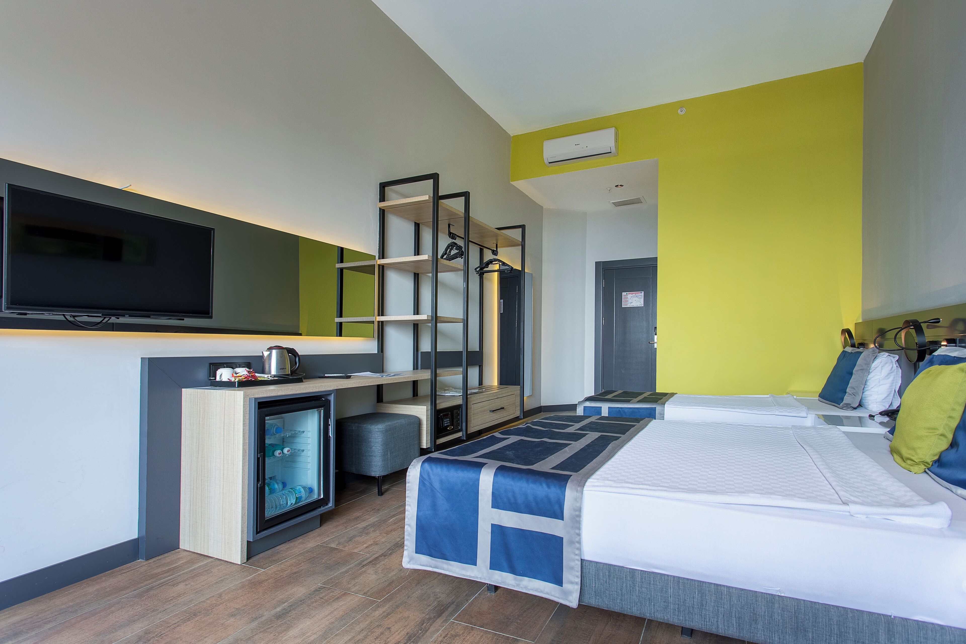 Ein modernes Hotelzimmer mit Flachbildfernseher, einer Minikühlschrank, einem Schreibtisch und zwei Betten mit blau-weißer Bettwäsche.