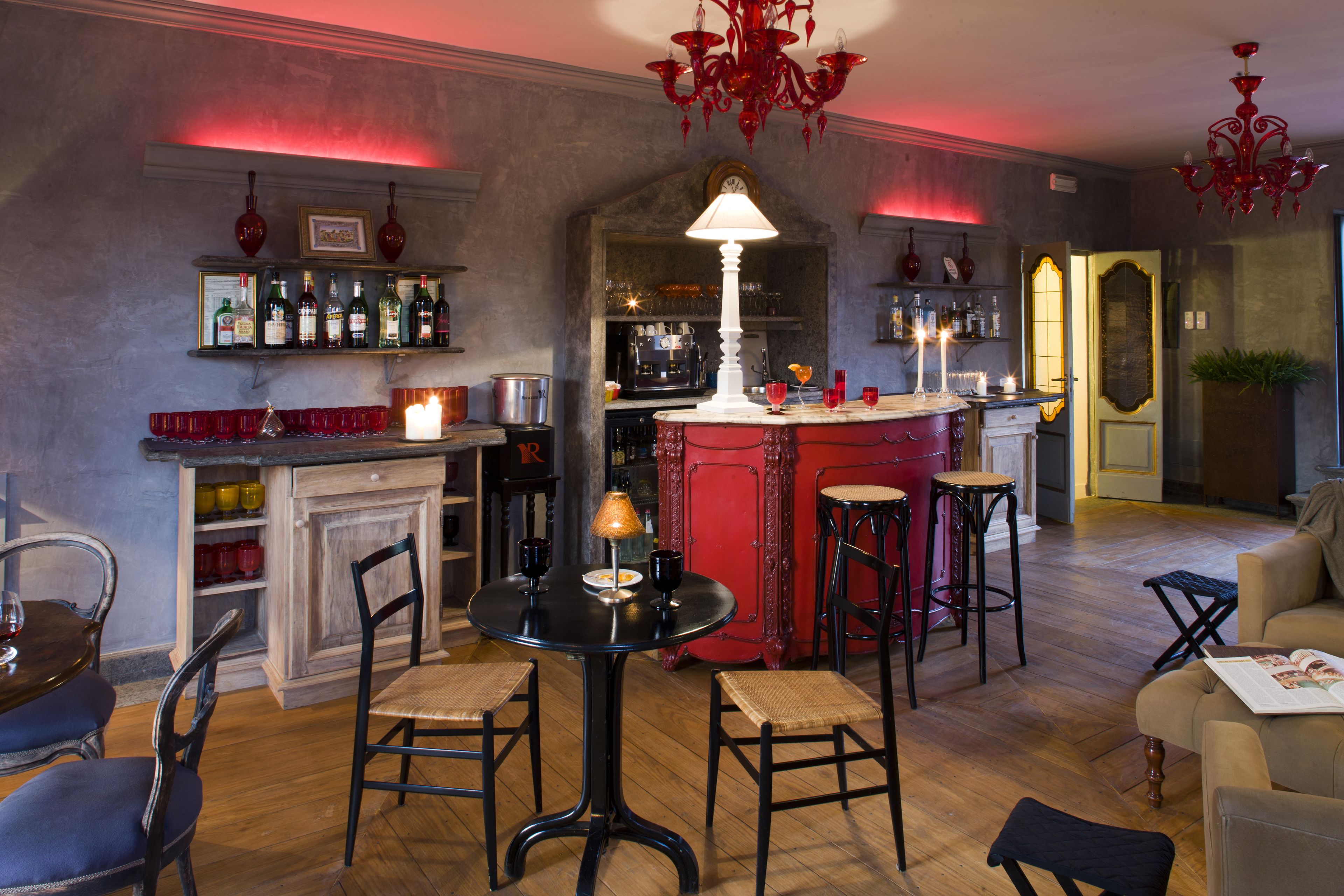 A cozy bar area with a round table, bar stools, and a red bar counter. There are shelves with bottles and a chandelier above.