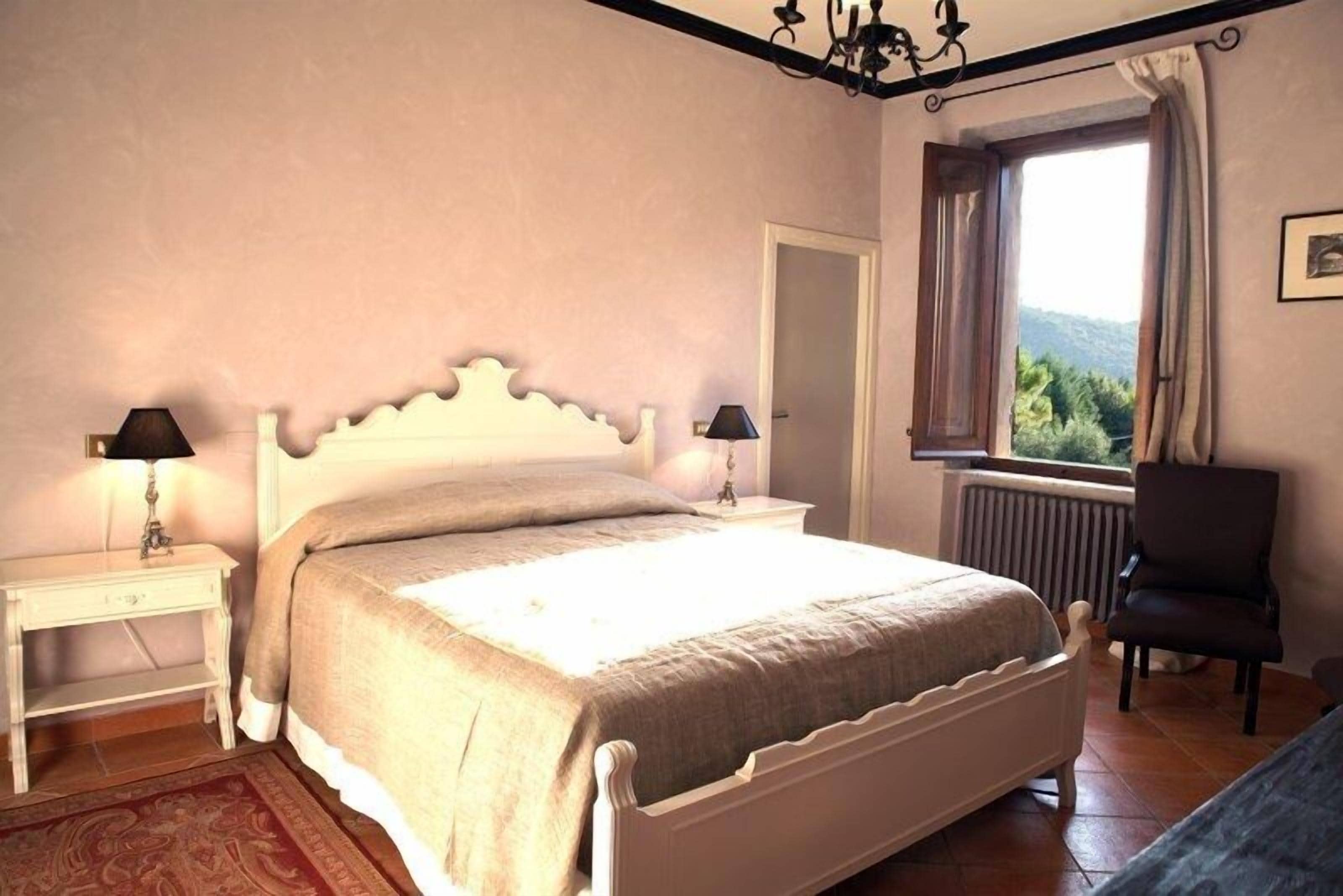 Quarto com cama, duas mesas de cabeceira com abajures, uma cadeira, janela com vista e radiador.