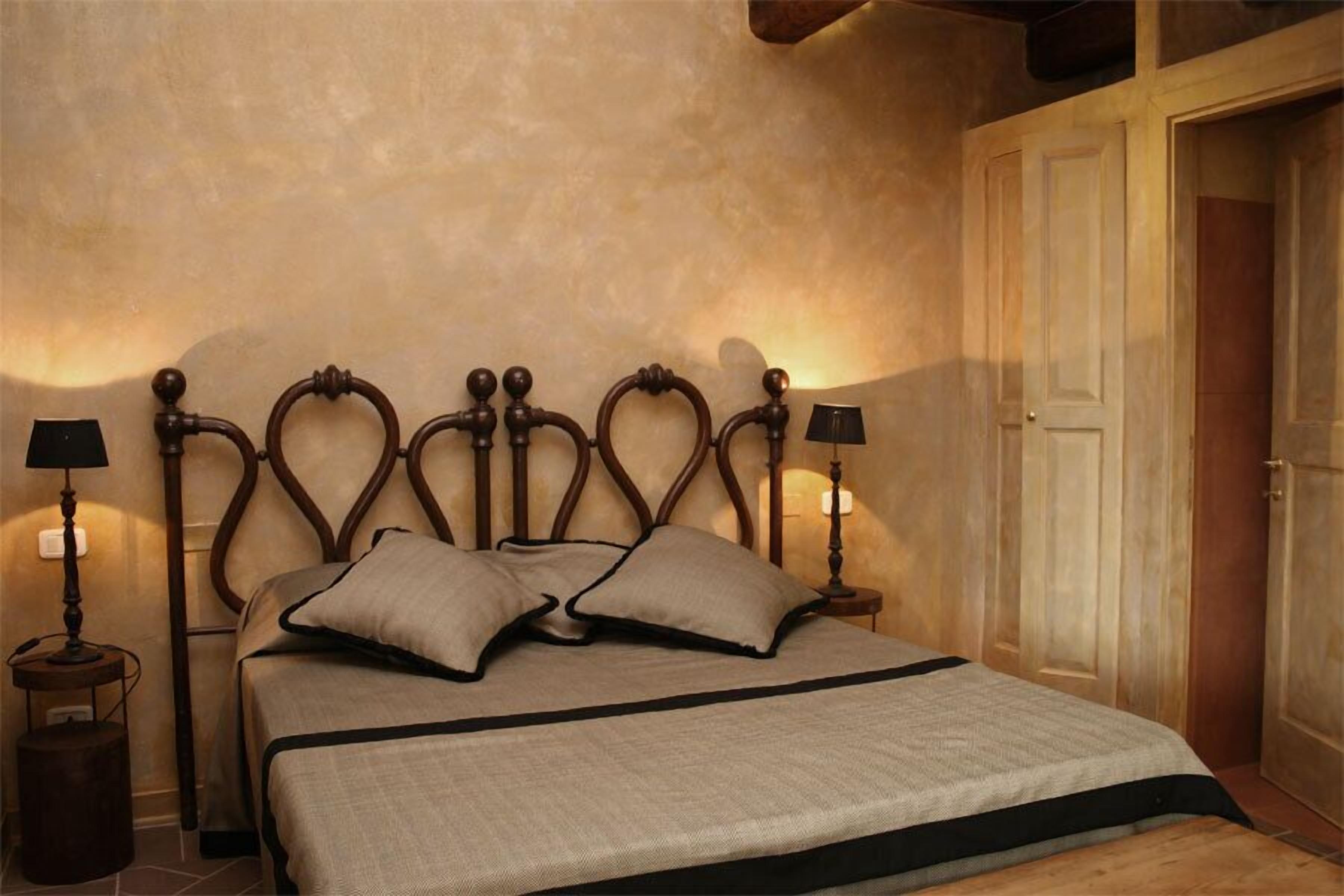 Une chambre avec une tête de lit en bois, deux lampes de chevet, un lit recouvert d’une couverture beige et, au fond, une porte en bois.