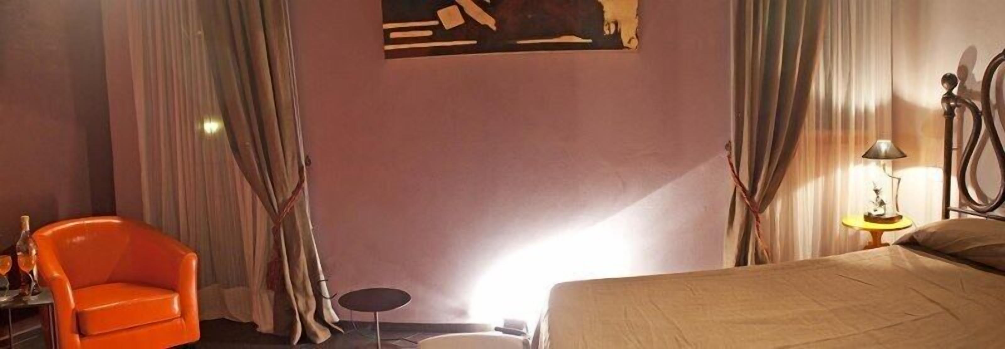 Una habitación de hotel con una cama, un sillón naranja, una mesita con una botella y una lámpara.