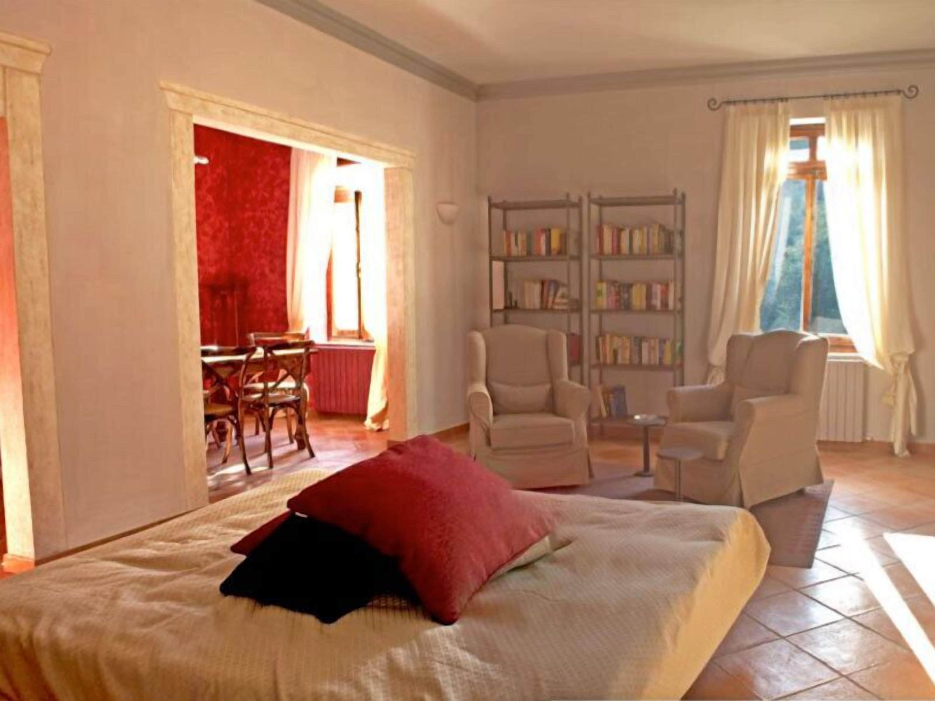 Un dormitorio con cama, una silla, una mesita, una estantería y una ventana con cortinas.