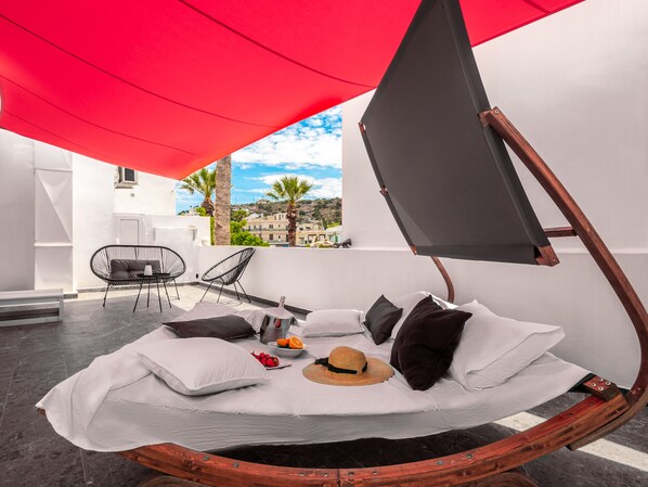 A modern outdoor lounge area with a hammock bed, a red canopy, and a view of the cityscape. - Kalypso Suites Hotel (Agios Nikolaos)