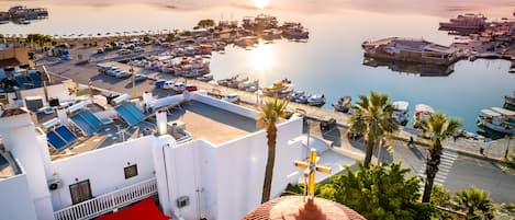 A marina with boats docked, a white building with a red awning, and a view of the sea and mountains.