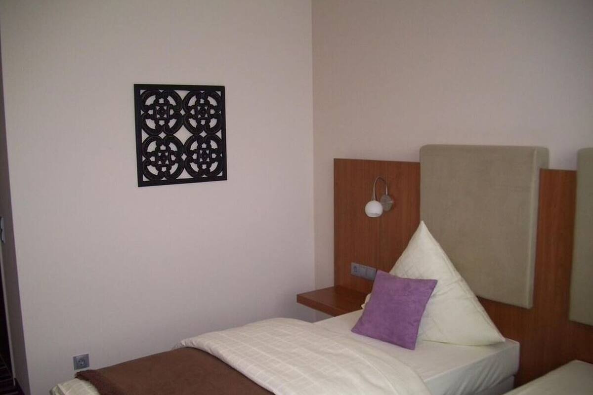 Una cama individual con una manta marrón, un almohadón blanco y un almohadón morado. Arriba de la cama, hay una lámpara fijada a la pared.