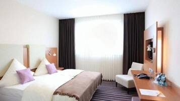 Una habitación de hotel con cama, escritorio, silla y un gran ventanal con cortinas.