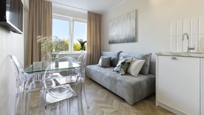 Een moderne woonkamer met een grijze bank, een glazen eettafel en stoelen, een witte kast, en een raam met gordijnen.