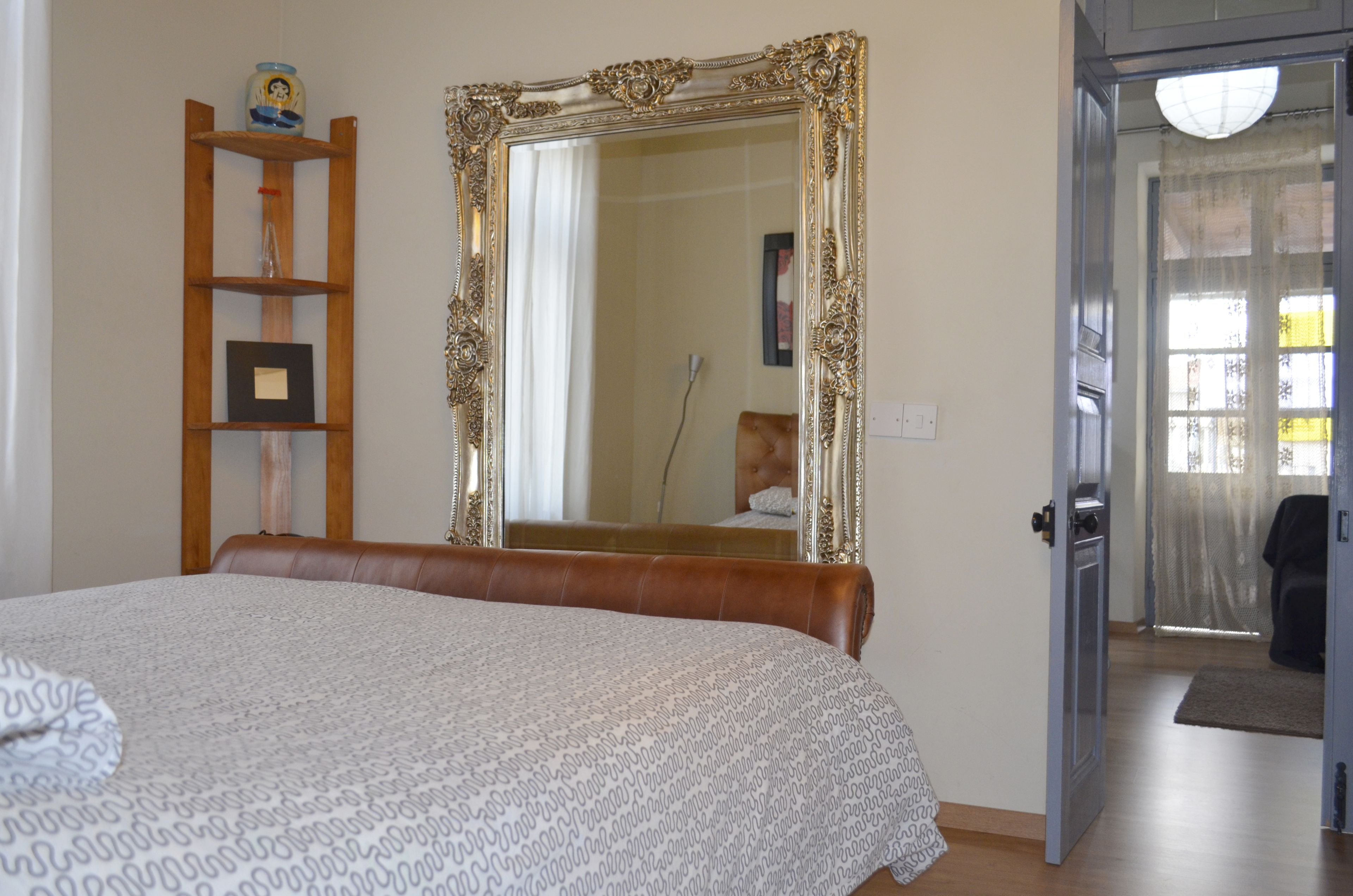 A bedroom with a large ornate mirror, a wooden shelf with a picture frame, and a bed with a patterned bedspread.