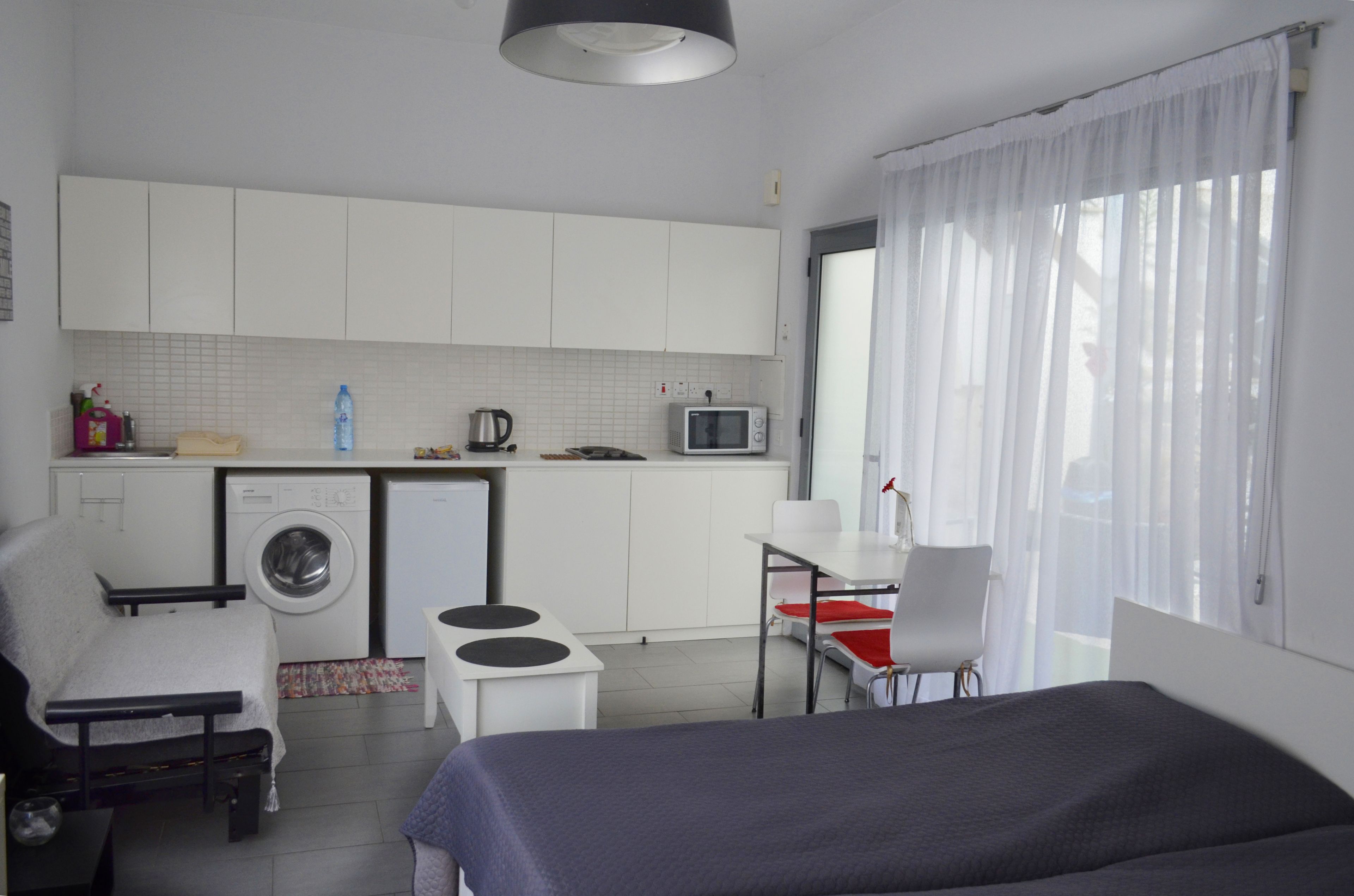 A compact hotel room with a bed, a small kitchenette, a washing machine, a dining area, and a window with curtains.