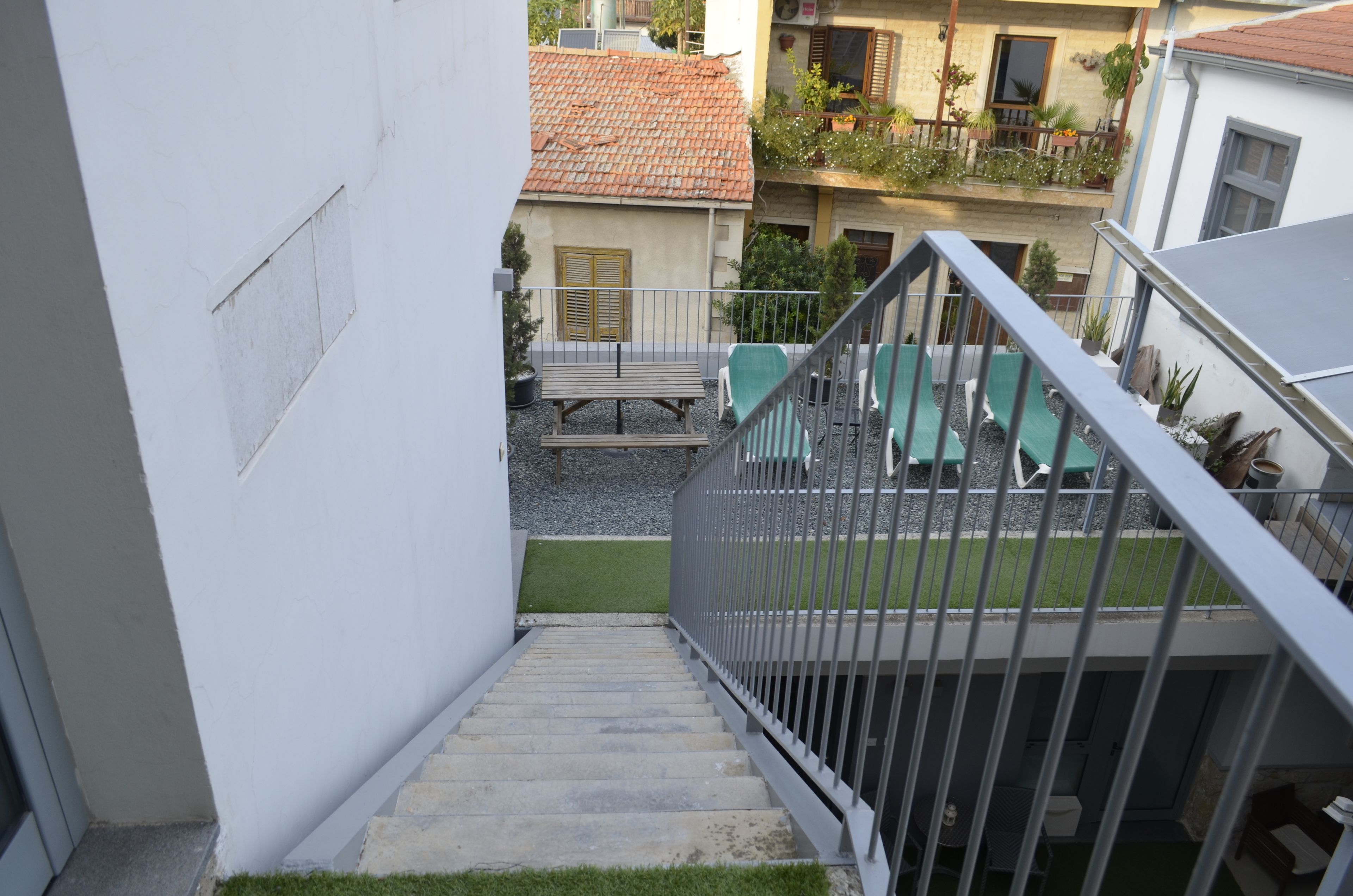 Escadaria que dá para uma varanda com mesa e cadeiras, cercada por uma parede branca e um edifício com telhado de azulejos.