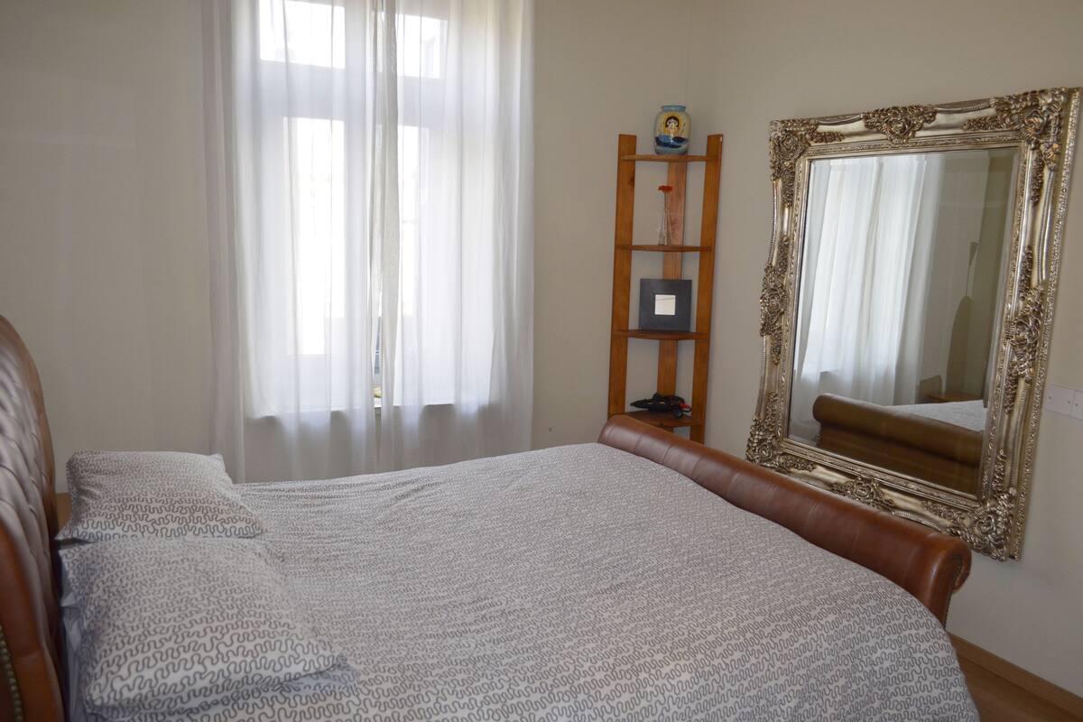 Un dormitorio con una cama grande, una estantería de madera tipo escalera, un espejo y una ventana con cortinas translúcidas.