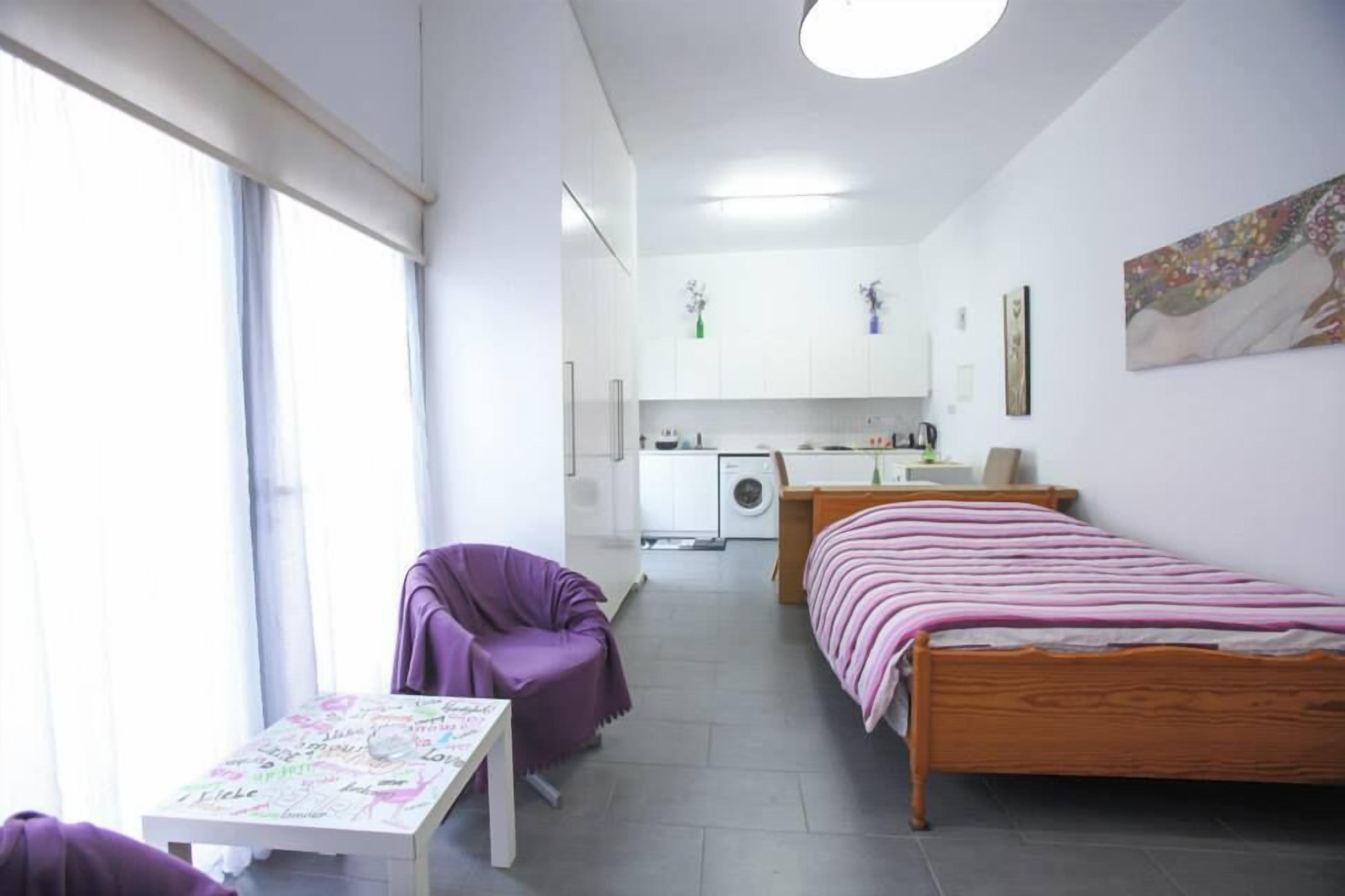 Una stanza compatta con un letto, un tavolino, una poltrona viola, angolo cottura e un'ampia finestra.