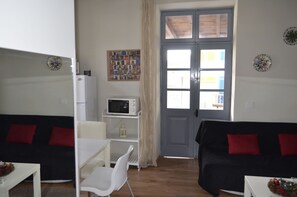 Un espacio habitable pequeño con un sofá negro, una mesa blanca, un microondas y una puerta con ventanal.